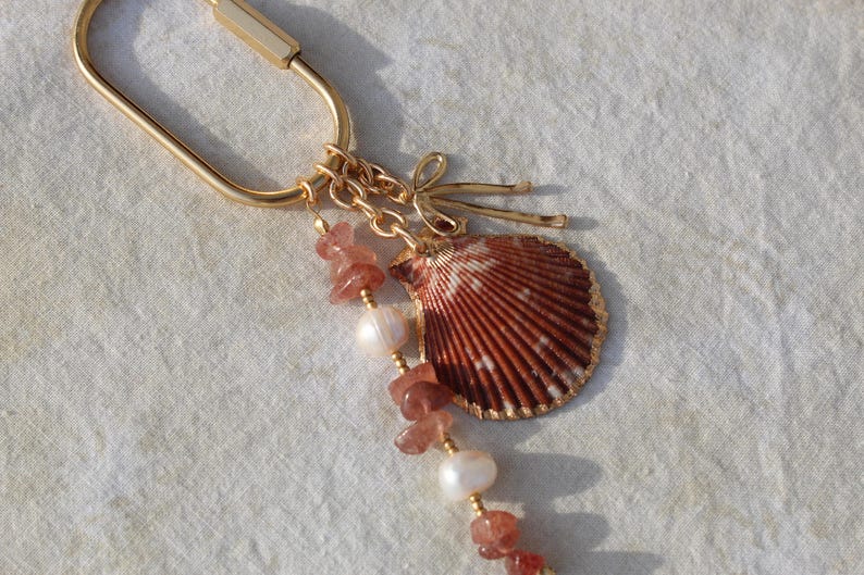 Angled view of full coastal bag charm keychain showing gold carabiner clasp, strawberry quartz chip beads, freshwater pearls and natural scallop shell pendant on linen