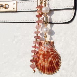 May include: A decorative bag charm featuring a strand of pink stone chips and a strand of white pearls and gold beads, finished with a large, fan-shaped, brown and white striped seashell pendant, attached to a white handbag with gold hardware.