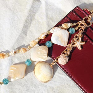May include: A gold-toned keychain with seashell and starfish charms, paired with a burgundy wallet. The keychain features a chain with a seashell and starfish charm, and a strand of seashells, square shells, and blue beads.
