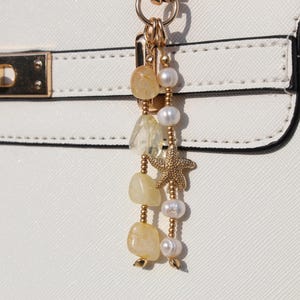 May include: A white handbag featuring a gold-toned metal clasp and a decorative hanging charm. The charm consists of two strands: one with yellow tumbled stones and small gold beads, and another with white pearls and a gold-toned starfish charm.