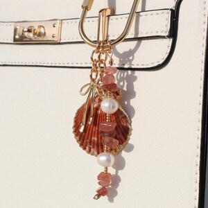 Strawberry quartz and pearl beaded bag charm keychain with scallop shell pendant and gold bow charm clipped to white structured handbag hardware