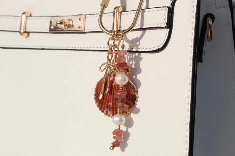 Strawberry quartz and pearl beaded bag charm keychain with scallop shell pendant and gold bow charm clipped to white structured handbag hardware