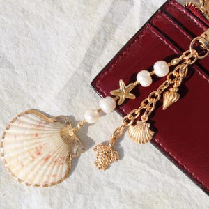 May include: A gold-toned keychain with a seashell charm, pearl beads, and other sea-themed charms. The keychain is attached to a burgundy leather cardholder, creating a coastal theme.