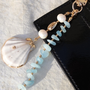 May include: A seashell charm with gold accents and a beaded lanyard featuring aquamarine stones, pearls, and gold beads. The lanyard is attached to a black leather wallet with a gold clasp.