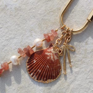 Coastal bag charm keychain with natural scallop shell pendant with gold dipped edges, strawberry quartz chip beads, freshwater pearls and gold bow charm on gold carabiner clasp on linen