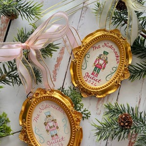 May include: Two oval-shaped gold-toned frames containing hand-painted nutcracker illustrations. Each frame features the text "The Nutcracker" and "Juliet 2026" in red script. The ornaments are adorned with sheer pink and white ribbons for hanging.