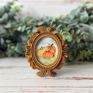 May include: A small, oval-shaped, gold-colored frame with an ornate design. Inside the frame is a watercolor painting of three pumpkins in shades of orange and yellow. The background is a light blue sky and green grass.