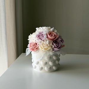 Könnte beinhalten: Ein Blumenarrangement mit pastellfarbenen Rosen in Rosa, Lavendel und Creme, akzentuiert durch weiße Füllblumen. Der Strauß befindet sich in einem weißen, zylindrischen Keramiktopf, der mit einem erhabenen Herzmuster verziert ist.