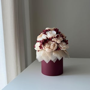 Könnte beinhalten: Eine zylindrische burgunderrote Geschenkbox mit einem Blumenarrangement aus cremefarbenen skulptierten Blüten, kleinen weißen Blumen und dunkelrotem Trockenlaub, akzentuiert durch eine Schicht aus halbtransparentem beigem Dekorpapier.