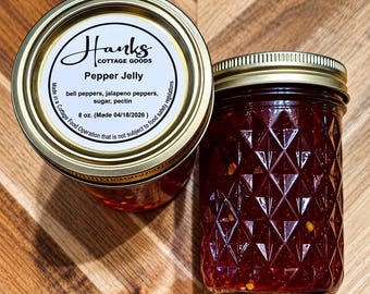 May include: Two glass jars of red pepper jelly with gold metal lids. The top label reads Hanks Cottage Goods, Pepper Jelly, bell peppers, jalapeno peppers, sugar, pectin, 8 oz. (Made 04/18/2026). The jars feature a diamond-patterned texture.