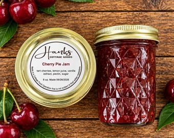 May include: A glass jar of red cherry pie jam with a gold lid and a label reading Hanks Cottage Goods, Cherry Pie Jam, 8 oz. Made 09/26/2026. The jar features a diamond-patterned texture and is surrounded by fresh red cherries and green leaves.