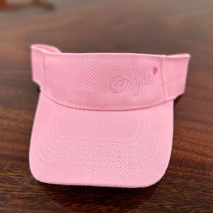 May include: A pink visor featuring the embroidered word "Alaska" in cursive, with a small pink heart. The visor is made of a textured material and has a curved peak to protect the eyes from sunlight.