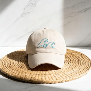 May include: A beige baseball cap with a curved brim, featuring an embroidered teal wave design on the front. The hat is displayed on a woven, circular mat, set against a marble background.