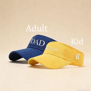 Könnte beinhalten: Zwei Sonnenblenden, eine blaue und eine gelbe, werden gezeigt. Die blaue Sonnenblende hat das Wort "DAD" in Weiß gestickt. Die gelbe Sonnenblende hat den Namen "Elias" und die Zahl "6" in Weiß gestickt. Die Wörter "Adult" und "Kid" sind ebenfalls sichtbar.