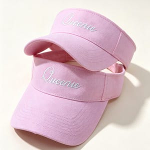 May include: Two pink visors stacked on top of each other. The word "Queenie" is embroidered in white on the front of each visor. The visors have a curved brim and a fabric band.