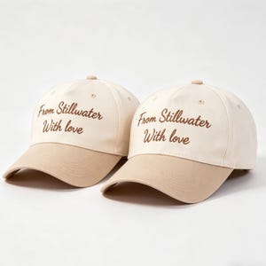 May include: Two baseball caps with a cream-colored crown and a tan brim. The caps have the text "From Stillwater With love" embroidered in brown script. The caps are a casual accessory.