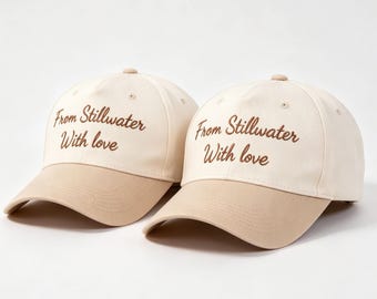 Könnte beinhalten: Zwei Baseballkappen mit cremefarbenem Oberteil und beigem Schirm. Auf den Kappen steht in brauner Schrift "From Stillwater With love". Ein lässiges Accessoire.