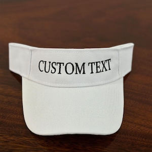 May include: White visor with the text "CUSTOM TEXT" embroidered in black. The visor is made of a soft, textured fabric and has a curved brim. The visor is designed to shield the eyes from the sun.