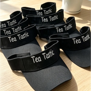Könnte beinhalten: Eine Ansammlung schwarzer Sonnenblenden mit dem weißen Schriftzug "Tea Tastic". Die Visore sind auf einer hellen Holzoberfläche angeordnet, was auf ein lässiges oder Outdoor-Thema hindeutet.