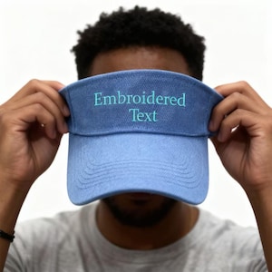 Puede incluir: Una visera azul claro con las palabras "Embroidered Text" bordadas en color turquesa. La visera está sujeta por una persona. La visera tiene una visera curva y una banda de tela.