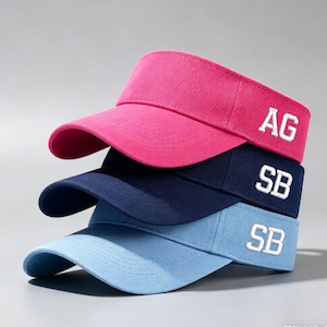 Op de afbeelding: Drie vizieren op elkaar gestapeld. Het bovenste vizier is felroze met de witte letters "AG". Het middelste vizier is marineblauw met wit "SB". Het onderste vizier is lichtblauw met wit "SB".