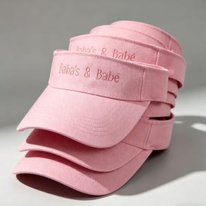Könnte beinhalten: Ein Stapel rosa Sonnenblenden. Jede Sonnenblende hat den gestickten Text "Baba's & Babe" in passendem rosa Garn. Die Sonnenblenden bestehen aus einem weichen, strukturierten Stoff und sind ordentlich gestapelt.