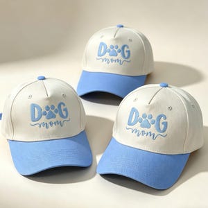 May include: Three baseball caps with a white crown and a light blue peak. The caps feature the embroidered text "DOG mom" in light blue, with a paw print design. The caps are a light, neutral colour.