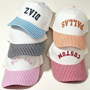 Op de afbeelding: Een collectie van vijf witte baseballpetten met geborduurde tekst en gestreepte kleppen. De petten bevatten de woorden DIAZ in donkerblauw, PALLAS in oranje, CUSTOM in rood en GUY MOM in lichtroze, gecombineerd met bijpassende blauwe, oranje, rode en roze strepen.