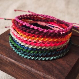Op de afbeelding: Een stapel van meerdere gevlochten koordarmbanden in verschillende kleuren, waaronder roze, rood, oranje, geel, blauw, groen en groenblauw. Elke armband heeft een gedraaid, geknoopt patroon en dunne, losse koorduiteinden voor verstelbaarheid.