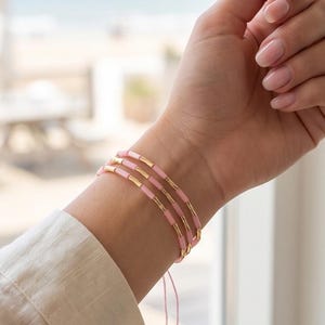 Könnte beinhalten: Ein dreireihiges Armband mit abwechselnden hellrosa zylindrischen Perlen und metallisch goldfarbenen Röhrenperlen. Das Armband wird mit einer dünnen rosa Kordel befestigt und hat zwei kleine hängende Perlen am Ende des verstellbaren Verschlusses.