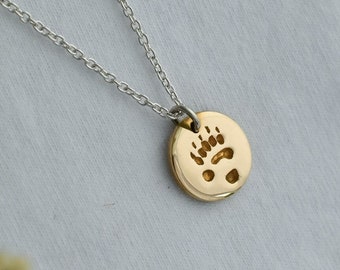 May include: A gold-colored pendant necklace with a silver chain. The pendant is a small, round disc with a bear paw print etched into it. The necklace is displayed on a light gray surface with dried flowers.