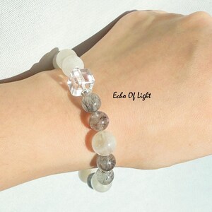 May include: A beaded bracelet featuring a mix of clear, gray, and white gemstones. The bracelet includes a faceted clear crystal and several round beads with inclusions. The text "Echo Of Light" is visible.