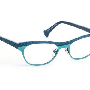 May include: Teal-colored eyeglasses with a cat-eye frame. The rims and temples are teal, with a textured pattern on the top of the frame and temples. The arms are a darker teal. The glasses have clear lenses and a nose bridge.