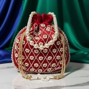 Może przedstawiać: Czerwona, okrągła, koralikowa torebka ze sznurkiem i perłową rączką. Torebka ma kwiatowy wzór i złoty sznurek. Tło jest z niebieskiej i zielonej tkaniny.