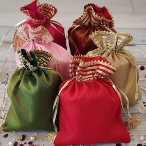 May include: An assortment of colorful drawstring pouches in shades of red, pink, green, gold, and brown. Each pouch is adorned with gold trim and tassels. The pouches are arranged on a light-colored wooden surface, with scattered decorative elements.