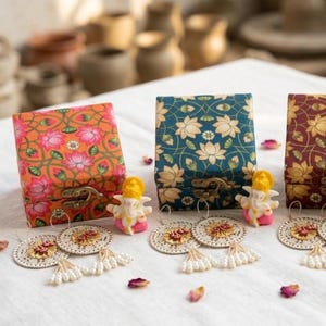 May include: Four square gift boxes with floral patterns in orange, teal, maroon, and green, each featuring a metal latch. Beside each box sits a small Ganesha figurine and a pair of circular, white, embroidered earrings with dangling white beads.