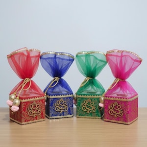 May include: Four colorful gift boxes with gold accents and a sheer fabric top. The boxes are red, blue, green, and pink, with gold tassels and a decorative design. They are arranged on a wooden surface.