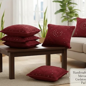 May include: A collection of five burgundy cushion covers with intricate mirror work detailing. The square pillows are arranged on a dark brown wooden table and a beige sofa. The cushions have a textured surface and a decorative pattern.