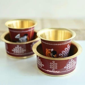 May include: Two nested maroon and gold-colored cups. The cups have a gold rim and a maroon body with white decorative patterns and a painted image. The cups are sitting on a white surface.