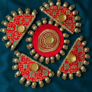 Op de afbeelding: Decoratieve rode en gouden stoffen stukken gerangschikt op een donkerblauwe achtergrond. Elk stuk heeft een goudkleurig metalen accent en een patroon met groene en witte details. Het centrale stuk is een rode cirkel met gouden versieringen.