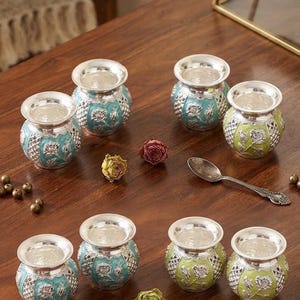 May include: A set of eight small, round metal pots with silver-toned rims and bases, featuring embossed floral patterns in turquoise and lime green enamel. The pots are arranged in two rows, accompanied by a silver spoon and several dried rosebuds.