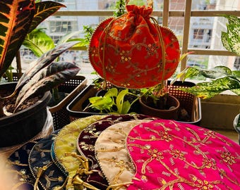 Könnte beinhalten: Eine orangefarbene, runde, bestickte Tasche mit einem goldperlenbesetzten Henkel hängt vor einer Sammlung ähnlicher Taschen. Die Taschen sind in verschiedenen Farben erhältlich, darunter Pink, Blau und Gelb, und sind mit goldener Stickerei und Quasten verziert.