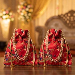 May include: Two red drawstring pouches featuring intricate floral embroidery in gold, blue, and green threads. Each bag is adorned with a double-strand pearl beaded handle and gold metallic trim along the edges, creating a structured silhouette.