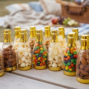 Könnte beinhalten: Eine Sammlung von durchsichtigen Kunststoffflaschen mit goldfarbenen Verschlüssen und Böden, die jeweils mit verschiedenen Snacks wie braunen Mandeln, weißem Popcorn und bunten Schokolinsen gefüllt und auf einer Holzoberfläche angeordnet sind.