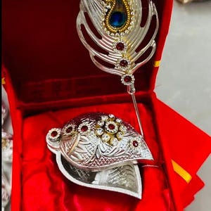 May include: A silver-colored decorative feather and headpiece set, adorned with red and clear gemstones, rests inside a red box. The feather features a blue and gold accent. The headpiece is intricately designed with floral patterns.