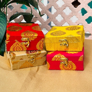 Può includere: Quattro piccole scatole decorative rosse, gialle, dorate e rosa. Ogni scatola ha una chiusura dorata ed è adornata con un motivo floreale dorato. Le scatole sono impilate, mostrando i loro colori vivaci e i loro intricati disegni.
