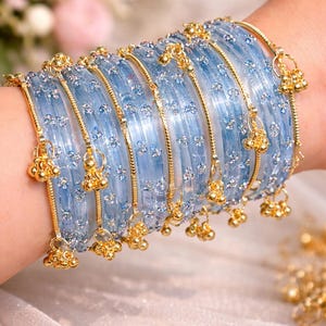 Puede incluir: Un conjunto de pulseras azul claro con detalles dorados y colgantes. Las pulseras están adornadas con pequeños detalles brillantes y se apilan en una muñeca. Los colgantes dorados añaden un toque de elegancia.