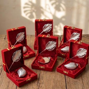 May include: Several red velvet boxes with gold trim are open, revealing silver feather-shaped ornaments and small silver containers. The boxes sit on a wooden surface, with the contents displayed prominently.