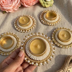 Puede incluir: Cinco candelabros decorativos con velas encendidas. Los candelabros son redondos, con una base de metal dorado, rodeados de perlas blancas y cuentas doradas. Las velas son amarillas. Flores rosas y verdes en el fondo.