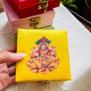 May include: Stack of decorative boxes in red, pink, and yellow. The top of each box features a gold clasp. The top box is red, the middle box is pink, and the bottom box is yellow. Each box has a printed image of a deity.